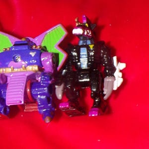 May include: Two toy robots, one purple and green with wings, the other black with a silver chest and a white arm.