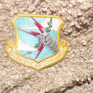 May include: A shield-shaped embroidered patch featuring a light blue sky with white clouds, a silver armored gauntlet grasping red lightning bolts, and a green olive branch. A yellow border surrounds the patch with the text STRATEGIC AIR COMMAND.