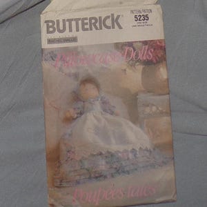 以下が含まれることがあります： ピローケースドール用のButterickソーイングパターン、パターン番号5235。 パターン封筒には、花のアクセントが付いた白いドレスを着た人形が描かれています。 テキスト「Pillowcase Dolls」が封筒の前面に表示されています。