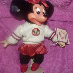 Puede incluir: Una mu&ntilde;eca de peluche Minnie Mouse roja y blanca que lleva una falda roja y un su&eacute;ter blanco con un logotipo de Mickey Mouse rojo. La mu&ntilde;eca est&aacute; de pie sobre un soporte blanco y tiene un lazo rojo en la cabeza. La mu&ntilde;eca sostiene una etiqueta que dice "Disney's" y "Minnie Mouse".