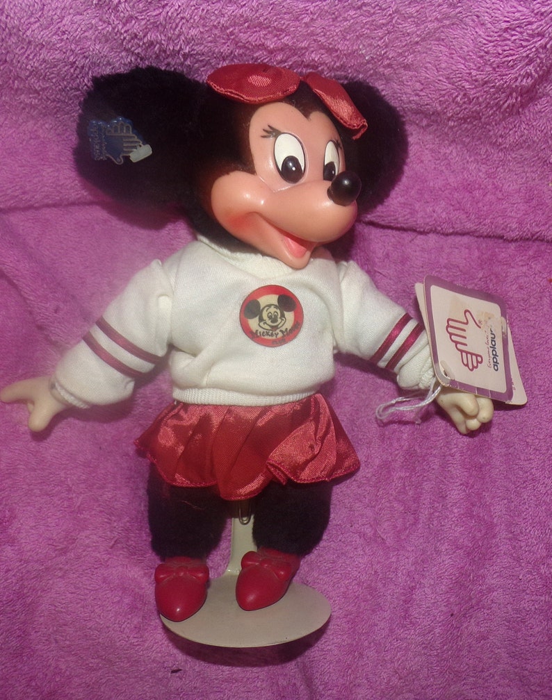 Puede incluir: Una mu&ntilde;eca de peluche Minnie Mouse roja y blanca que lleva una falda roja y un su&eacute;ter blanco con un logotipo de Mickey Mouse rojo. La mu&ntilde;eca est&aacute; de pie sobre un soporte blanco y tiene un lazo rojo en la cabeza. La mu&ntilde;eca sostiene una etiqueta que dice "Disney's" y "Minnie Mouse".