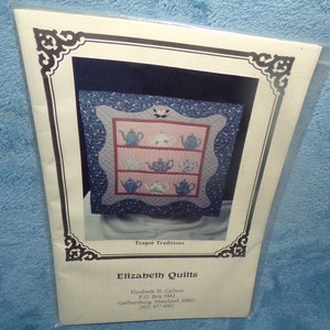 Könnte beinhalten: Ein verpacktes Quiltmuster mit einem gerahmten Design von Teekannen auf Regalen. Der Quilt hat ein rosa und blaues Farbschema mit floralen Akzenten. Die Verpackung enthält den Text "Teapot Traditions" und "Elizabeth Quilts".