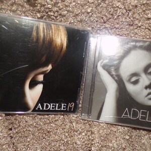 May include: Two music CDs in clear plastic cases. The left case features a dark portrait with the text ADELE 19. The right case displays a black and white portrait of a person with their hand near their head, featuring the text ADELE 21.