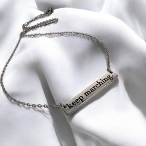 May include: A silver necklace with a silver bar pendant that says "keep marching."