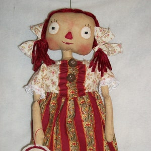 May include: A handmade rag doll with red yarn hair, a white and brown floral print dress, and a red and white striped skirt. The doll is holding a round tag that says "Hugs and Kisses".