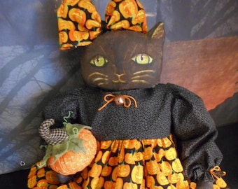 Puede incluir: Una muñeca de tela hecha a mano con forma de gato negro de ojos verdes, que viste una blusa negra de manga larga y una falda con estampado de calabazas naranjas. El gato lleva un lazo a juego en la cabeza y sostiene una pequeña calabaza de tela.