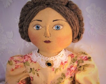 May include: A fabric doll with brown yarn hair styled in a braided crown, blue eyes, and painted facial features. The doll wears a pale yellow dress with a pink rose floral pattern, puff sleeves, and a white lace collar with a small black brooch.