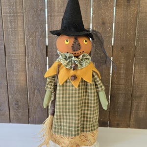 May include: A decorative Halloween scarecrow doll with a pumpkin head, wearing a black witch hat and a plaid dress. The doll has a yellow collar and holds a small broom. The dress is green and yellow plaid with a lace trim.