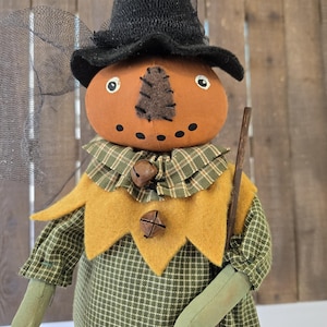 May include: A handcrafted scarecrow doll with a pumpkin-shaped head, black hat, and green plaid shirt. The doll has a yellow collar with bells and holds a wooden stick. A black net is attached to the hat. A festive autumn decoration.
