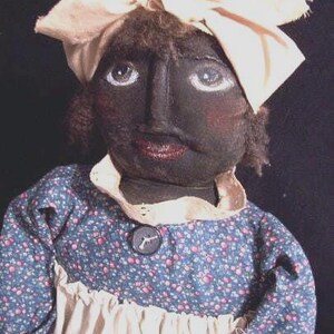 May include: A handmade doll with a dark complexion, wearing a blue floral dress with a white apron and a cream-colored headscarf tied in a bow. The doll has painted facial features, including large eyes and red lips. A button adorns the dress.