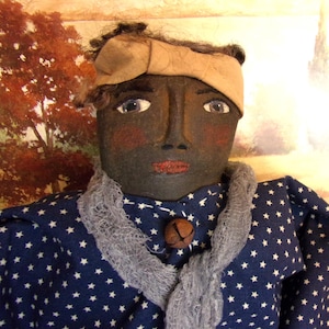 May include: A handmade doll with a dark complexion, wearing a blue dress with white stars, a gray scarf, and a tan headband. The doll has painted facial features, including rosy cheeks and red lips. A small bell is attached to the dress.