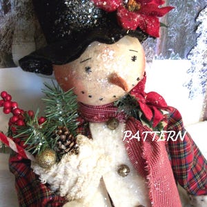 May include: A handcrafted snowman figure with a black top hat, red poinsettia, and a red plaid scarf. The snowman has a carrot nose, black dot eyes, and a white coat with gold bells. It holds a sprig of pine, berries, and a candy cane. The word "PATTERN" is visible.