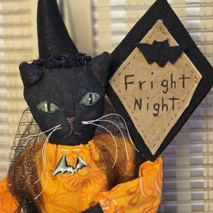May include: A black cat figurine wearing a pointed black hat and an orange floral patterned shirt. The cat has green eyes, white whiskers, and a silver bat pin. It holds a black diamond-shaped sign with a tan center that reads Fright Night.