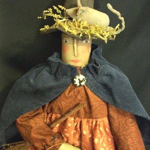 May include: A handcrafted witch doll with a painted face, wearing a blue cape and an orange dress. The doll has a straw hat adorned with yellow sprigs and a small pumpkin. It holds a wooden stick and a broom.