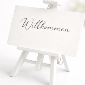 Könnte beinhalten: Ein kleiner weißer Staffelei-Ständer hält eine weiße Karte mit dem Wort "Willkommen" in eleganter Schrift. Die Staffelei ist aus Holz und hat drei Beine. Der Hintergrund ist ein weiches, verschwommenes Weiß.