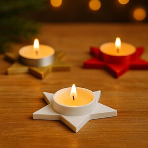 May include: Three lit tea light candles on star-shaped holders. One holder is white, one is gold, and one is red. The candles are burning with a warm glow, set against a blurred background of lights and greenery. The holders are on a wooden surface.