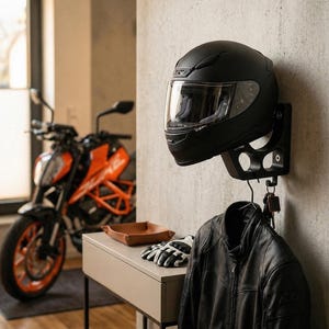 Può includere: Un casco da moto nero montato su una staffa a muro, con visiera trasparente. Sotto, una giacca di pelle nera è appesa a un gancio con chiavi. Sullo sfondo si vede una moto, guanti e un piccolo vassoio.