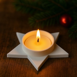 May include: A lit candle in a white ceramic holder shaped like a star. The candle is in a round, white container. The background includes a Christmas tree branch and a red ornament. The candle flame is bright.