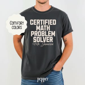 May include: Dark grey t-shirt with the text "CERTIFIED MATH PROBLEM SOLVER Mr. Jameson" in white. The shirt also has the words "Comfort Colors" in a white cloud-shaped design. The word "pepper" is at the bottom of the shirt.