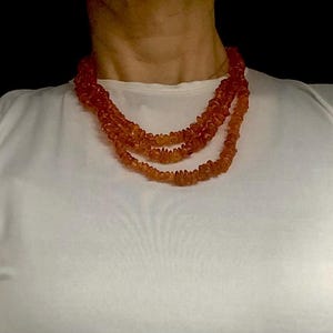 May include: A multi-strand necklace made of amber beads. The beads are a warm, honey colour and are arranged in a graduated pattern, with the largest beads at the centre of the necklace. The necklace is worn by a person with a white shirt.