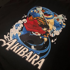 Puede incluir: Camiseta negra con un estampado gráfico de un personaje del anime "Demon Slayer" montando una criatura azul similar a una tortuga. El texto "AKIBARA" está impreso en blanco en la parte inferior de la camiseta.