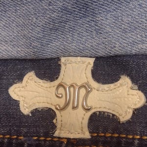 May include: Close-up of denim fabric with a white leather patch featuring a silver "M" design. The patch is in the shape of a cross. The denim is a medium wash blue.