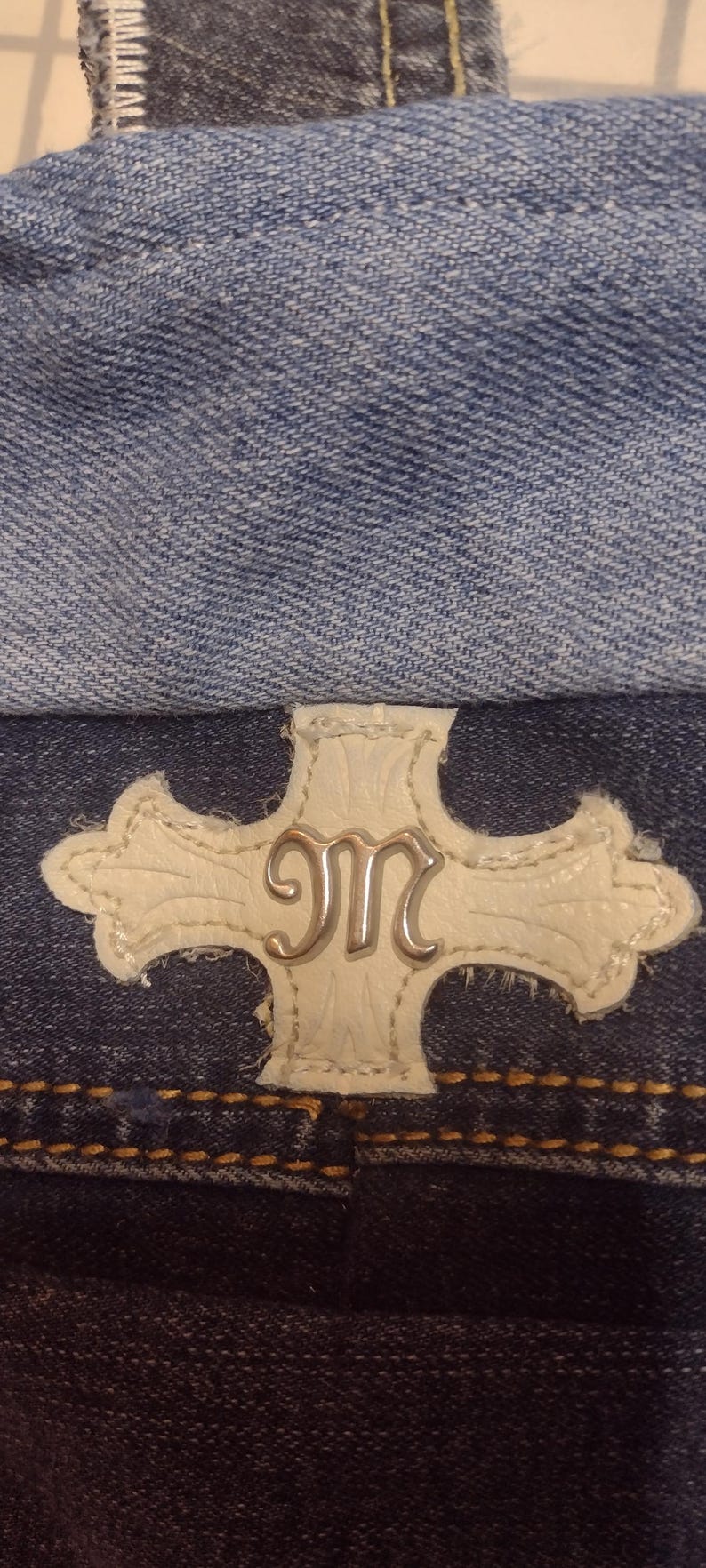 May include: Close-up of denim fabric with a white leather patch featuring a silver "M" design. The patch is in the shape of a cross. The denim is a medium wash blue.