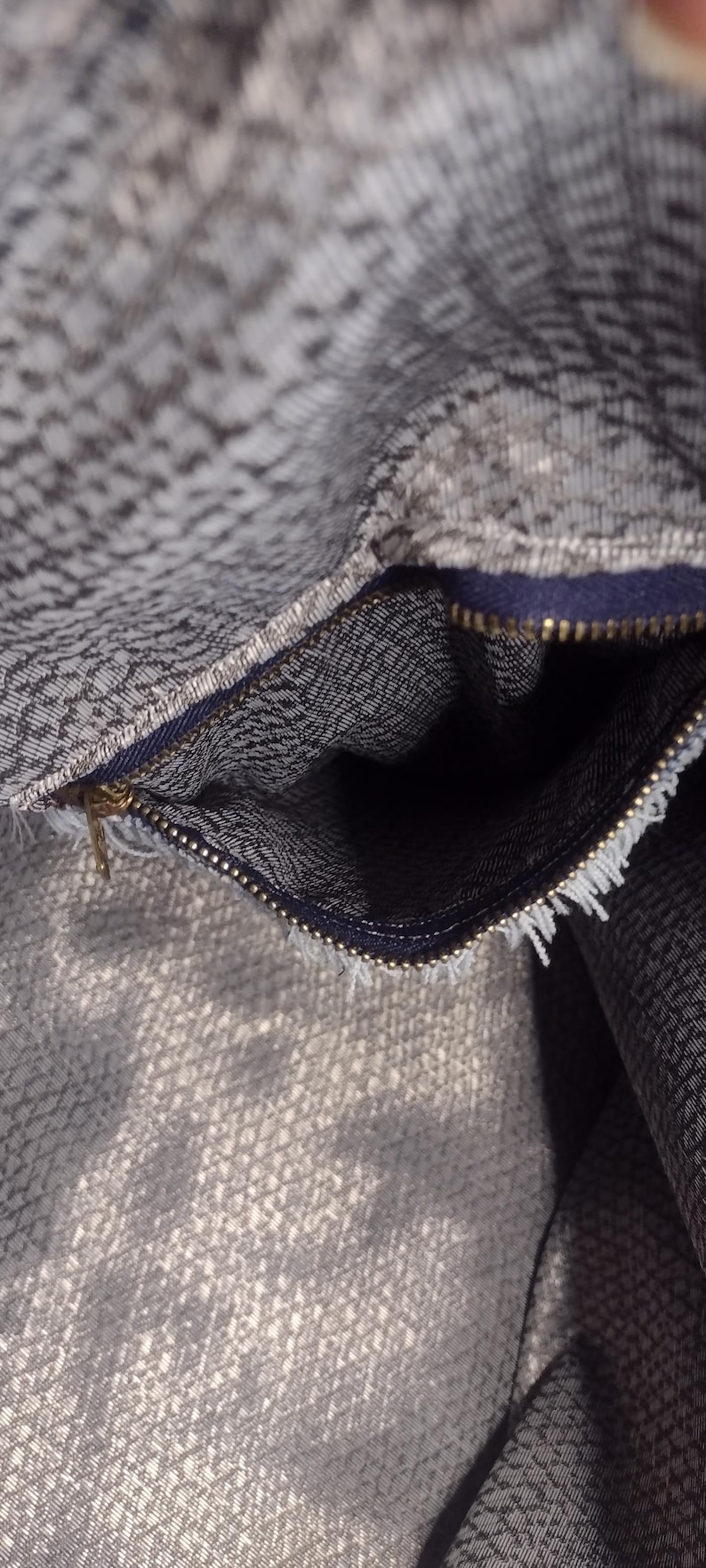 May include: Close-up of a gray and white patterned fabric with a zipper. The zipper is gold-colored and the fabric has a textured appearance. The interior of the fabric is visible.