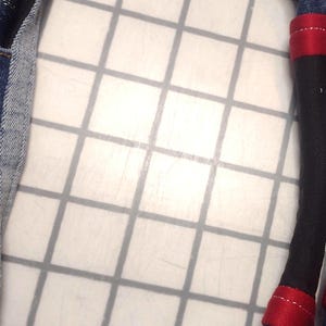 May include: Close-up of a denim fabric with a black fabric insert. Two black fabric legs with red accents and denim cuffs are visible. The legs are on a white grid background.