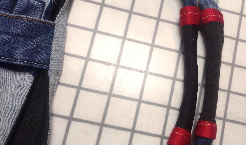 May include: Close-up of a denim fabric with a black fabric insert. Two black fabric legs with red accents and denim cuffs are visible. The legs are on a white grid background.