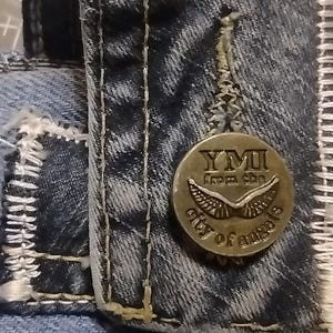 May include: Close-up of a denim jacket detail. A circular metal button with the text "YMI from the city of angels" is attached to the jacket. The jacket has white stitching and a light blue color.