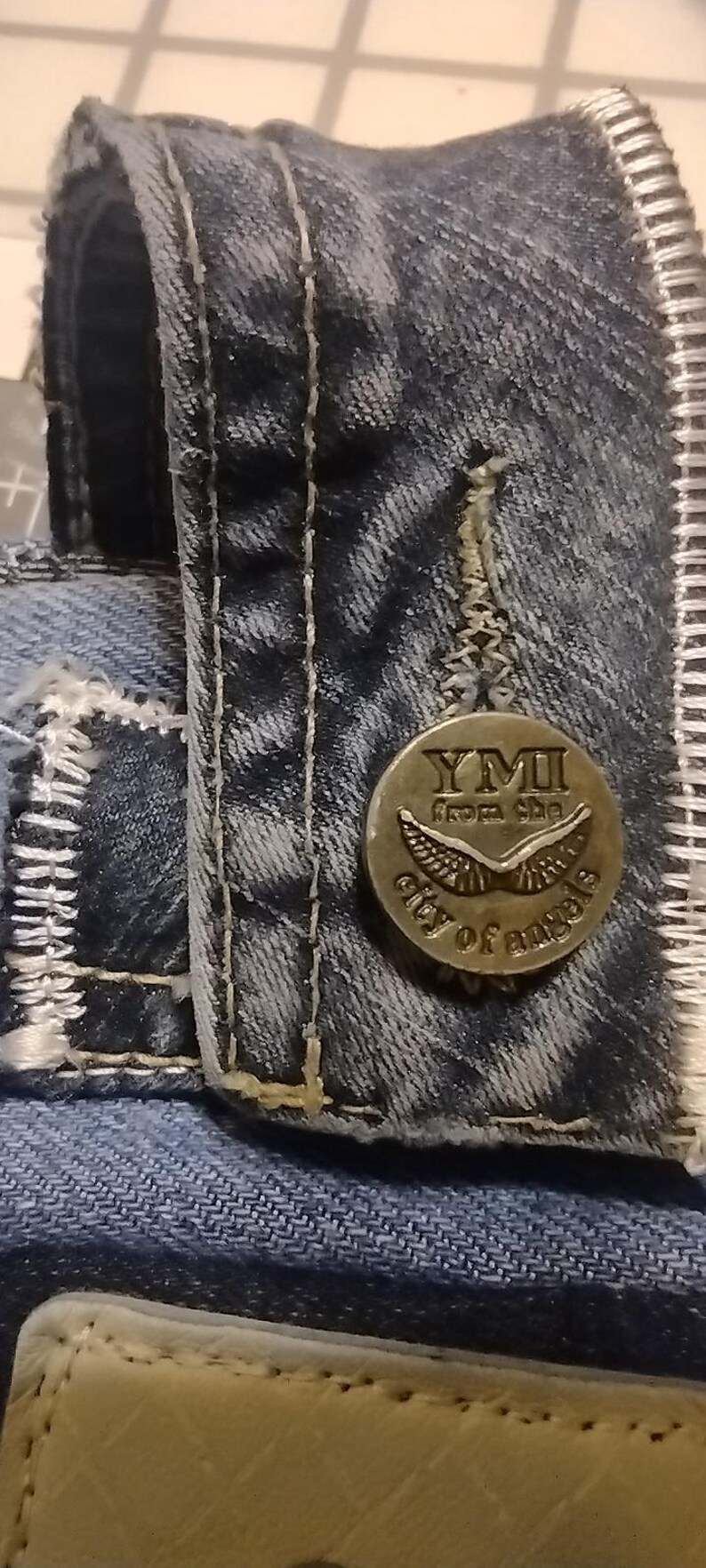May include: Close-up of a denim jacket detail. A circular metal button with the text "YMI from the city of angels" is attached to the jacket. The jacket has white stitching and a light blue color.