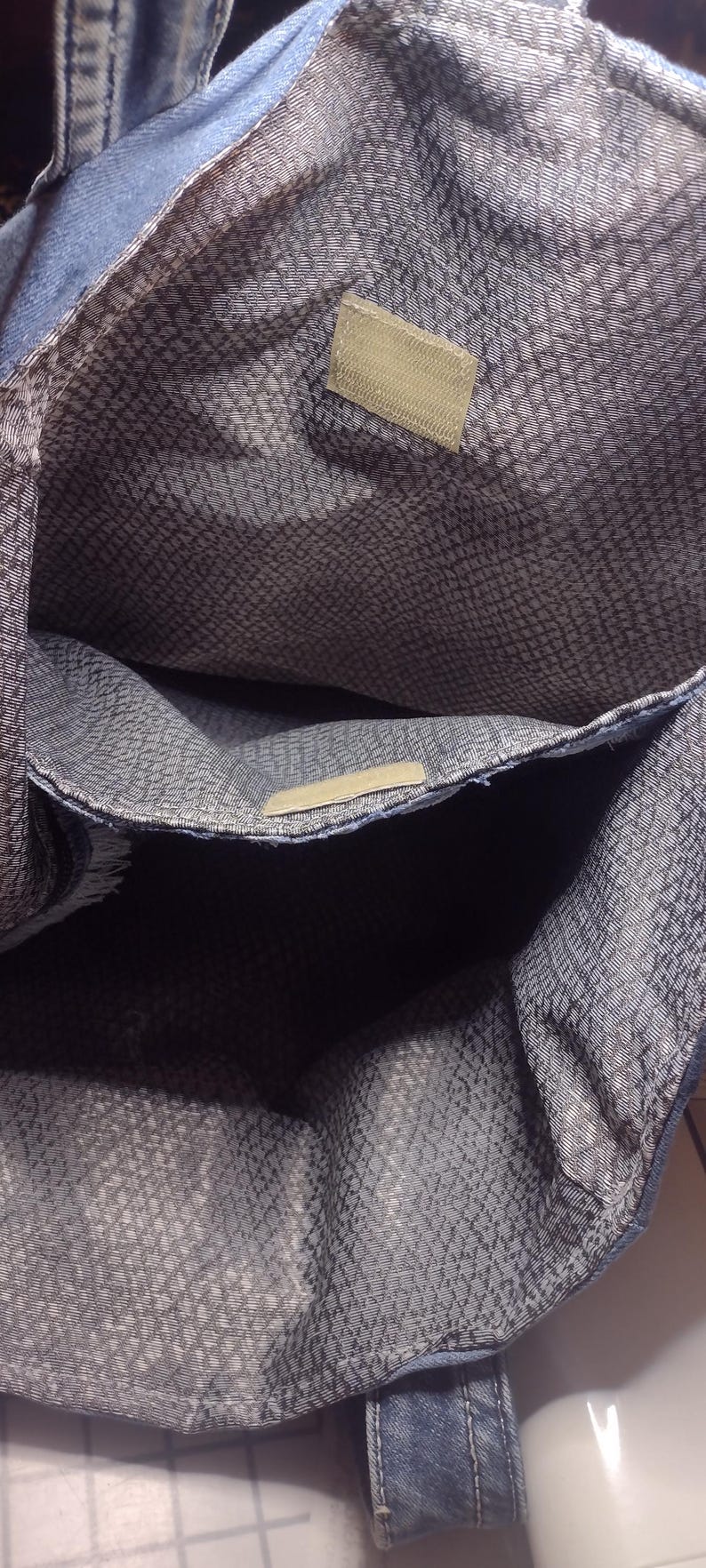 May include: A denim tote bag with a patterned interior. The bag has a rectangular shape with a large main compartment and a smaller pocket. The interior fabric has a gray and white pattern. The bag has a light blue denim exterior.