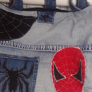 May include: Denim tote bag with a Spider-Man theme. Features a black spiderweb design, a spider pocket, and a red Spider-Man mask applique. The bag has a denim handle with red accents.