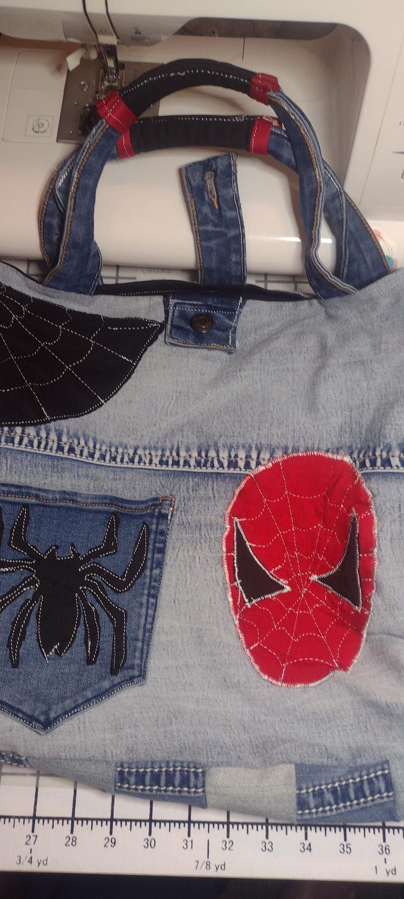 May include: Denim tote bag with a Spider-Man theme. Features a black spiderweb design, a spider pocket, and a red Spider-Man mask applique. The bag has a denim handle with red accents.