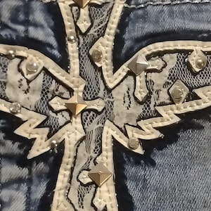 May include: Close-up of a denim garment with a large, ornate cross applique. The cross is cream-colored with silver studs and a lace overlay. The denim is a faded blue.