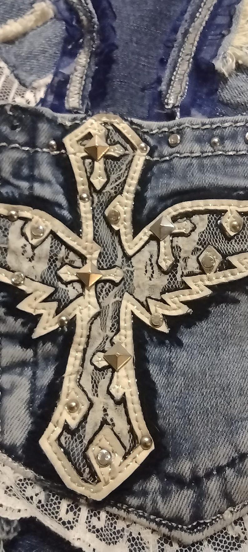 May include: Close-up of a denim garment with a large, ornate cross applique. The cross is cream-colored with silver studs and a lace overlay. The denim is a faded blue.