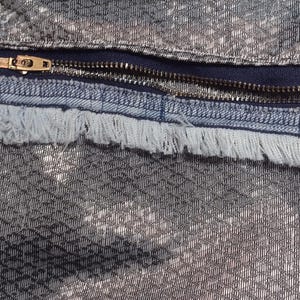 May include: Close-up of a textured fabric with a silver and black pattern. A dark blue zipper with a gold-colored pull is visible, along with a frayed edge of the fabric. The fabric has a woven appearance.