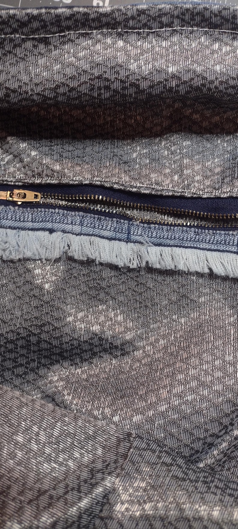 May include: Close-up of a textured fabric with a silver and black pattern. A dark blue zipper with a gold-colored pull is visible, along with a frayed edge of the fabric. The fabric has a woven appearance.