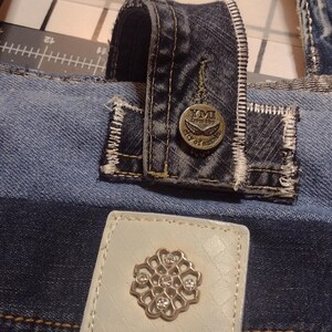 May include: Close-up of a denim tote bag with a white leather patch featuring a silver floral design and the text "Miss Me." The bag has a denim handle and a button with the text "YMI from the city of angels."