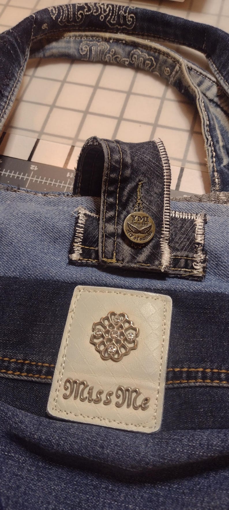 May include: Close-up of a denim tote bag with a white leather patch featuring a silver floral design and the text "Miss Me." The bag has a denim handle and a button with the text "YMI from the city of angels."