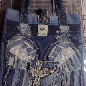 May include: Denim tote bag with a patchwork design. Features a pocket with a cross and wing design, lace accents, and the brand name "Miss Me". The bag has denim handles and a button closure.