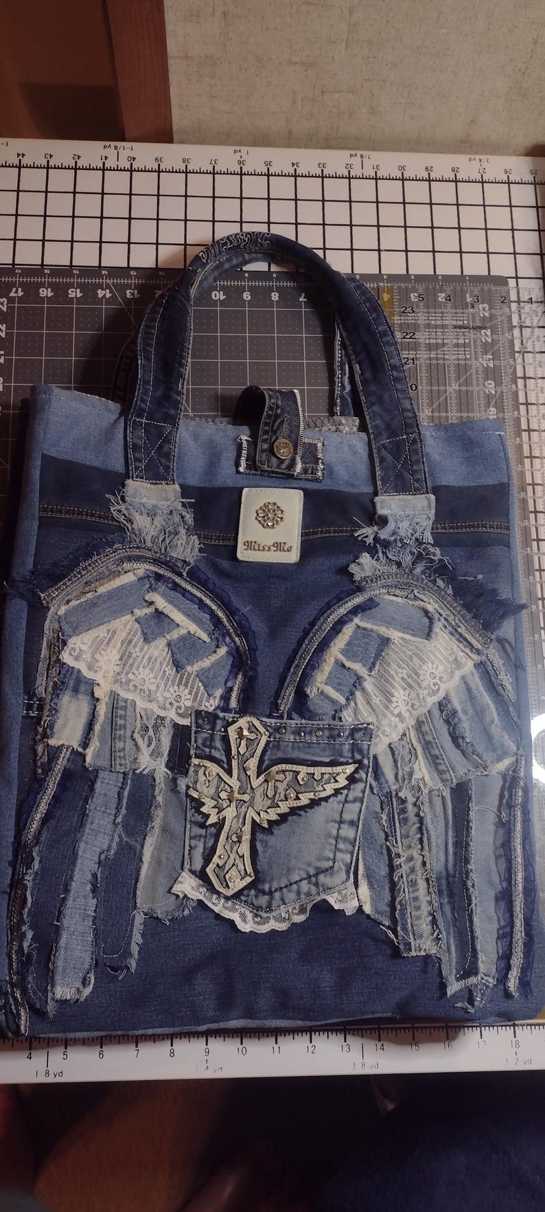 May include: Denim tote bag with a patchwork design. Features a pocket with a cross and wing design, lace accents, and the brand name "Miss Me". The bag has denim handles and a button closure.