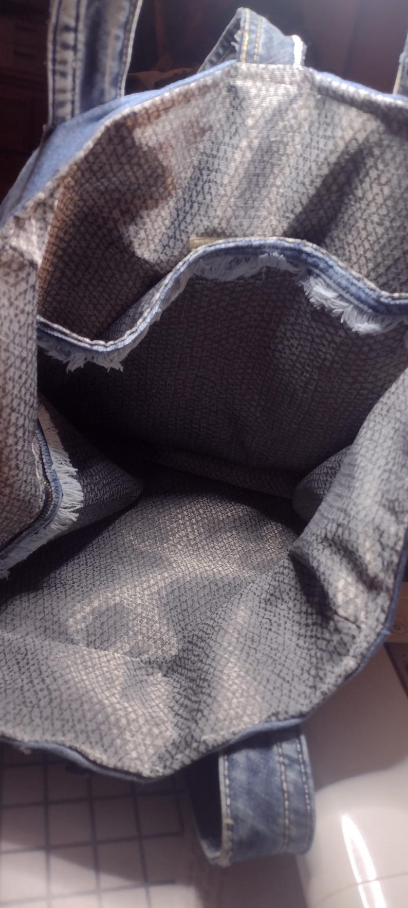 May include: A denim tote bag with a textured gray interior. The bag has frayed edges and denim handles. The interior fabric has a subtle pattern. The bag is open, showing the inside.