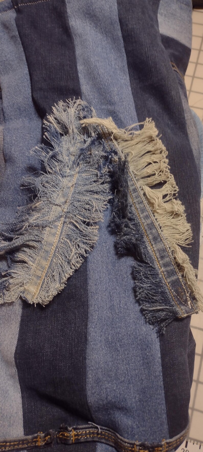 May include: Close-up of denim fabric with a patchwork design. Two fringed denim pieces are laid over the patchwork, showcasing frayed edges and a mix of light and dark blue hues. The overall texture is rough and casual.