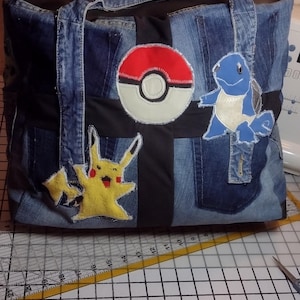 May include: Denim tote bag with embroidered Pokémon characters Pikachu and Squirtle, and a Poké Ball. The bag features a black panel and a zipper closure. The bag is on a cutting mat with measurements in inches.