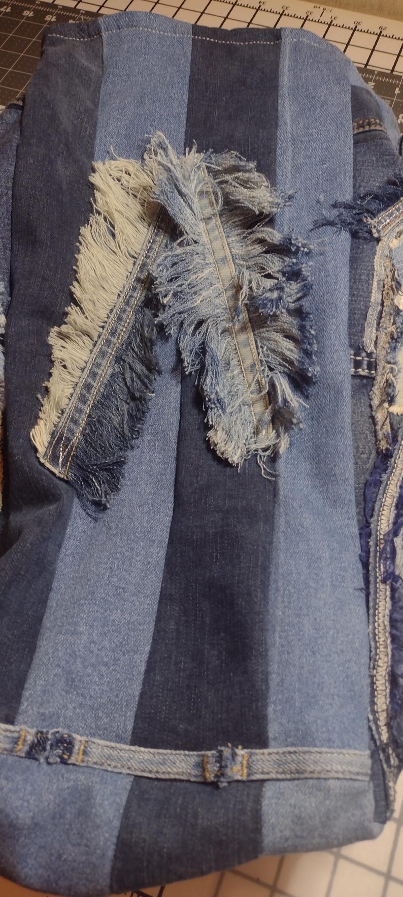 May include: Close-up of a denim textile with a patchwork design. The fabric features various shades of blue denim, with frayed edges and a striped pattern. The design includes a decorative fringe.
