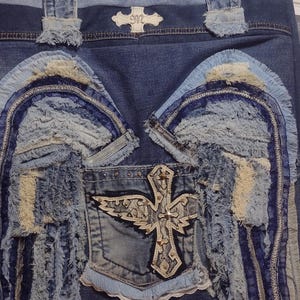 May include: Denim tote bag with a patchwork design. Features frayed fabric wings and a cross emblem on the front pocket. The bag has denim handles and a decorative emblem on the top.