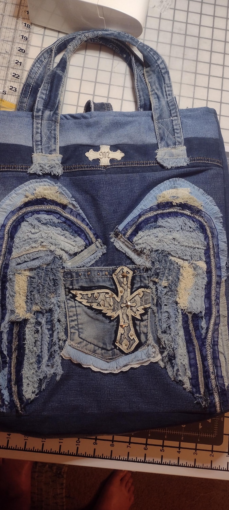 May include: Denim tote bag with a patchwork design. Features frayed fabric wings and a cross emblem on the front pocket. The bag has denim handles and a decorative emblem on the top.