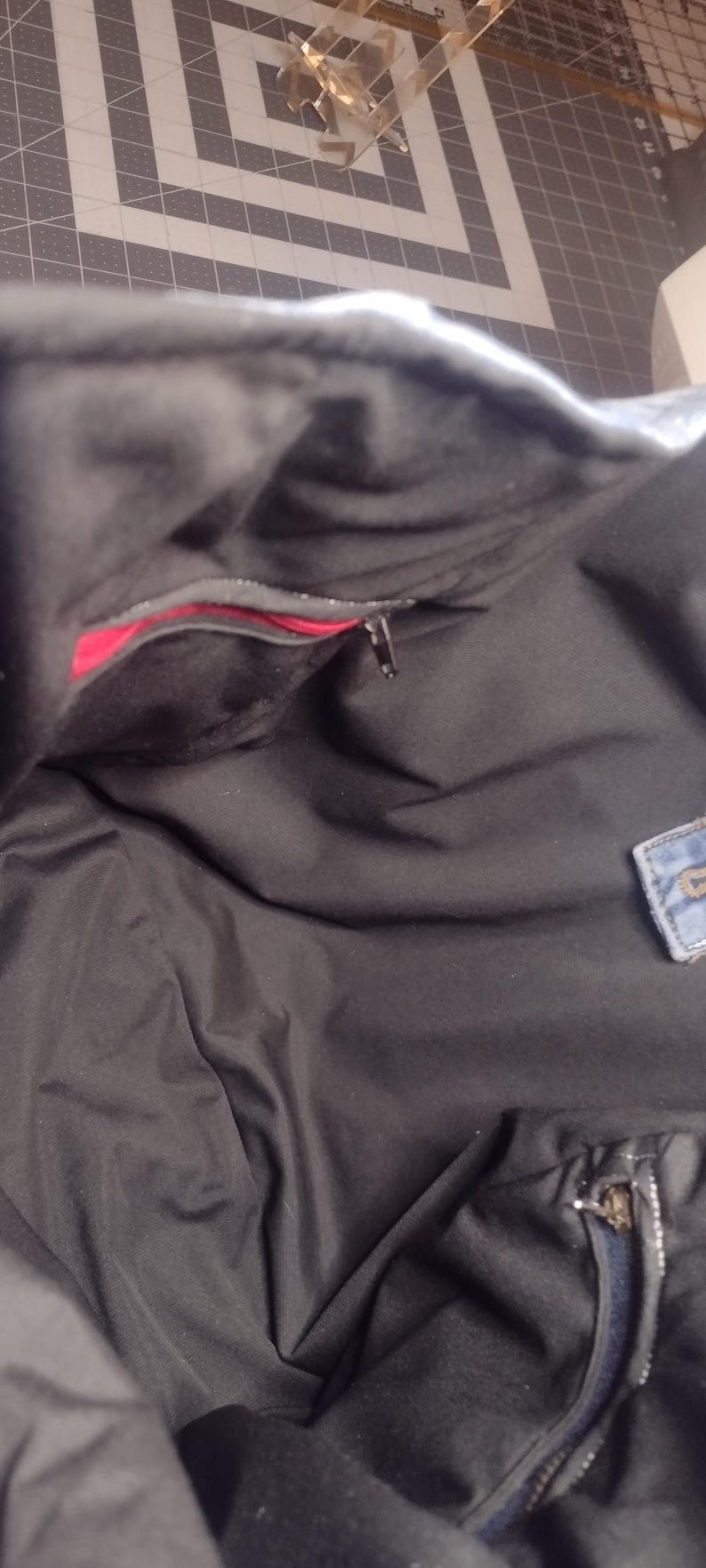 May include: Black tote bag interior with a red pocket and a zipper closure. The bag appears to be made of a soft, dark fabric. A small denim patch is visible on the side.